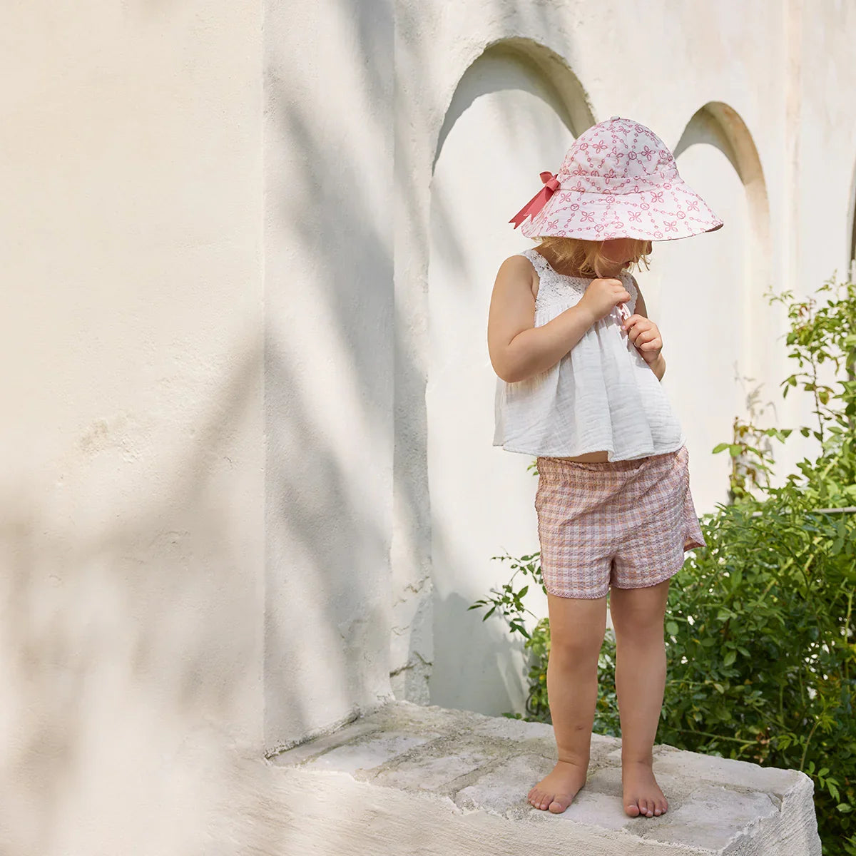 Deklica v belem poletnem klobuku s cvetličnim vzorcem, svetli majici in kratkih hlačkah stoji ob svetli steni na vrtu.